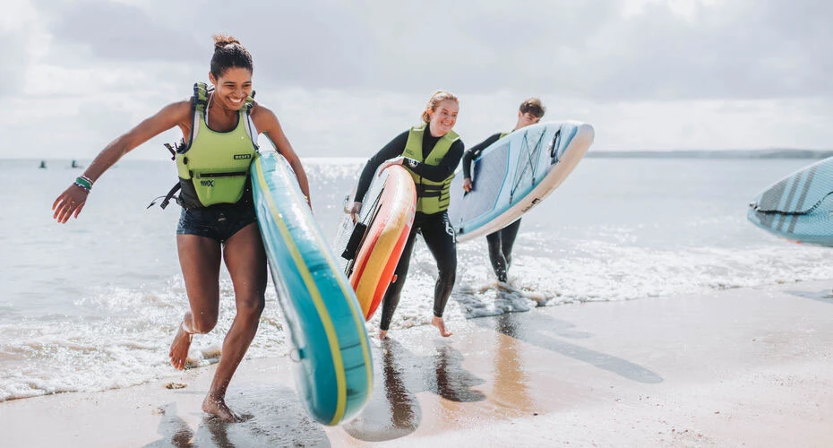 A group of students running out of the sea holding paddle boards