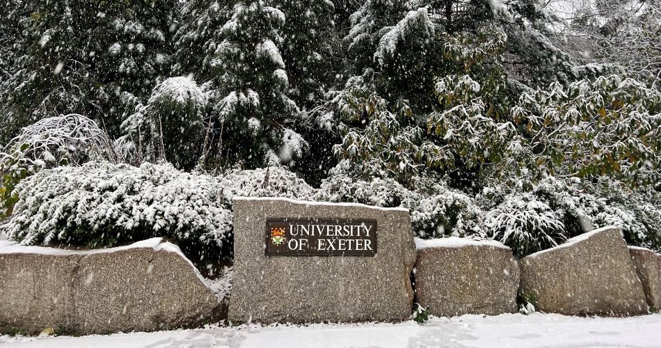 Large stones with a sign reading University of Exeter, in the snow against a backdrop of fir trees