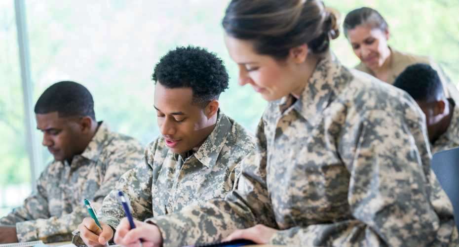 Three people dressed in camouflage combat attire sat at desks holding pens to write with