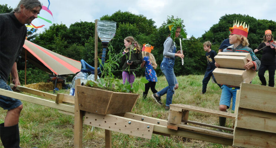 People participating in an outdoor activity involving a wooden structure that resembles a seesaw with plants and a water bottle attached. Children wearing colorful crowns and costumes are engaged in various activities, while adults assist and observe. The scene is set in a grassy area with trees in the background, suggesting a festive or community event.