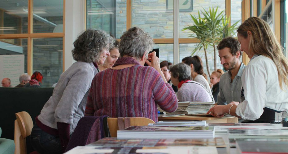 A group of people gathered around tables in a bright, indoor space with large windows and plants. They are engaged in various activities, such as reading, discussing, and examining materials. The atmosphere is collaborative and educational, suggesting a workshop or group learning environment. The participants are of various ages, contributing to a diverse and dynamic setting.