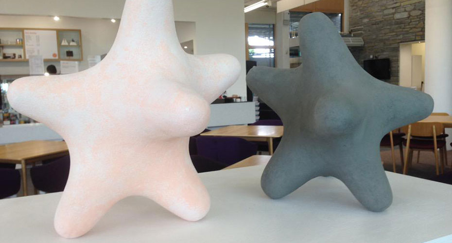 Two abstract sculptures by Samantha Gerlach resembling starfish with rounded edges are displayed on a white surface. One sculpture is a light peach color, while the other is dark gray. The background shows an indoor setting with tables, chairs, and a stone wall, suggesting the sculptures are part of an art exhibit in a public space.