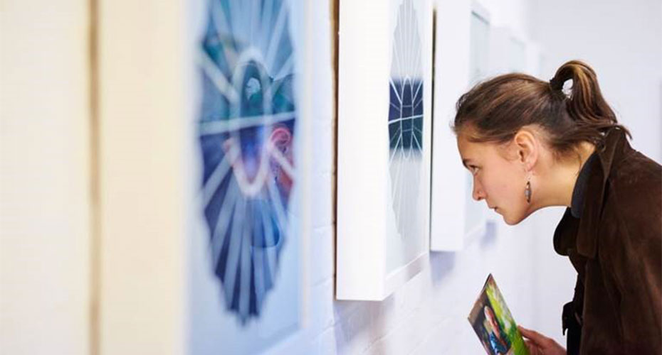 A woman with a ponytail closely examining a piece of art in a gallery. She holds a pamphlet in her hand and is intently looking at a framed artwork that features a geometric, starburst pattern. The gallery wall has multiple similar pieces displayed, creating a focused and contemplative atmosphere.