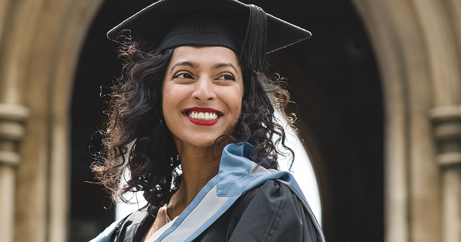 A young woman wearing a graduation cap and gown with a light blue sash stands outdoors, smiling brightly. She has curly dark hair and red lipstick. The background features an arched architectural structure, suggesting a university or similar institution.