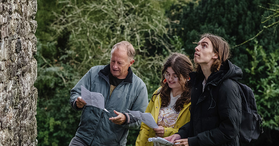 A group of three people outdoors, holding papers and examining a stone wall. One person, an older man in a blue jacket, looks intently at the paper. A young woman in glasses and a yellow jacket smiles at the document she holds. A young man with long hair and a black jacket gazes up thoughtfully. They are surrounded by greenery.