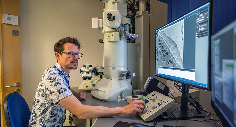 A man in a lab, examining a microscope and working on a computer.