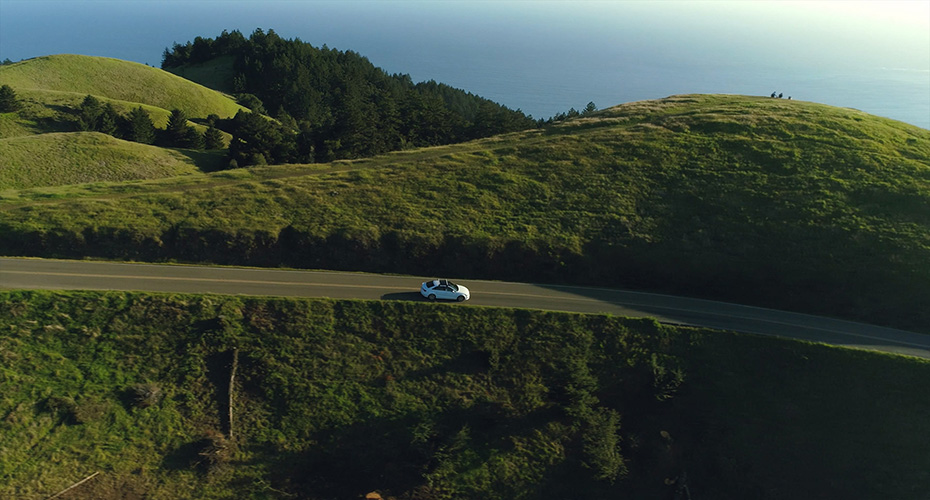Aerial view of car driving down country road through rural rolling hills with ocean in background at sunset, feels serene.
