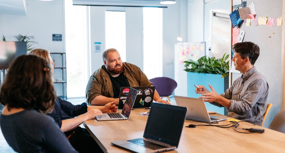 A diverse group of people sitting around a table with laptops, engaged in a meeting with graduates to discuss their business ideas.