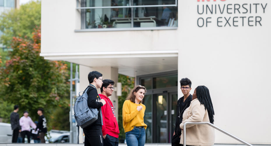 A diverse group of students engaged in discussion on the University of Exeter campus, surrounded by greenery and buildings.