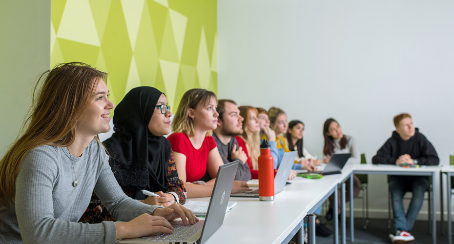 A diverse group of students collaborates at tables, each using laptops in a vibrant lecture space.