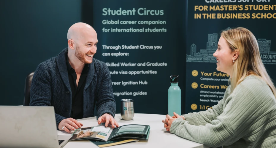 two people sat at a desk with a laptop and some brochures. In the background is a sign with text relating to careers support