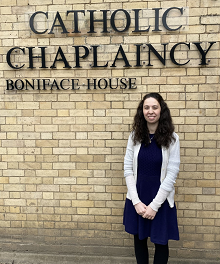 Maria Giannone, wearing a dark blue dress and white cardigan, standing in front of a light beige brick wall, which has dark green lettering attached to it. The lettering reads: 