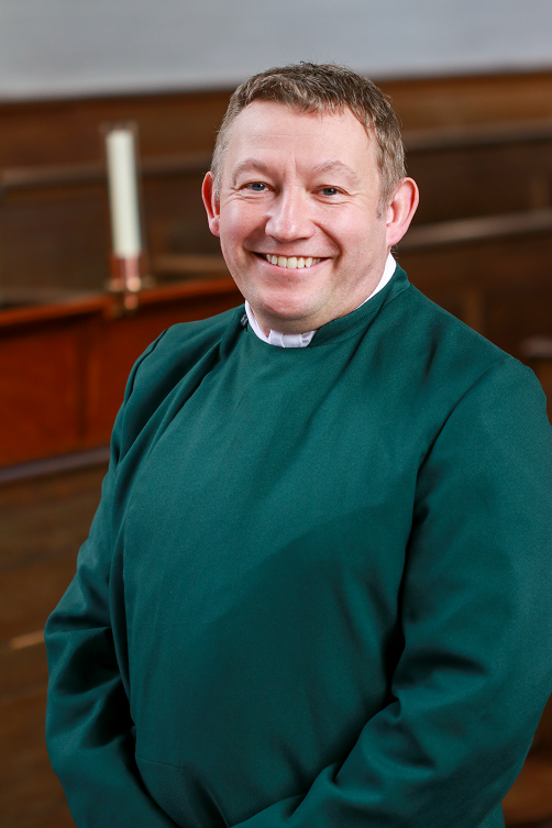 David Davies smiling at the camera while dressed in a double-breasted green cassock with a step collar opening showing a white cravat-style collar underneath. He is standing in front of wooden choir stalls, where one unlit white candle can be seen. Those who are familiar with the building would recognise this setting as the Mary Harris Memorial Chapel.