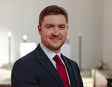 A photograph of Mr Michael Graham. He is wearing a red tie, white shirt and a dark blue suit, and is smiling at the camera. He is standing in the Mary Harris Memorial Chapel. The white east wall and altar, draped in a white altar-cloth, is softly focused behind him in the distance. The altar is partially sunlit, with silver-grey candlesticks gleaming in the sunlight.