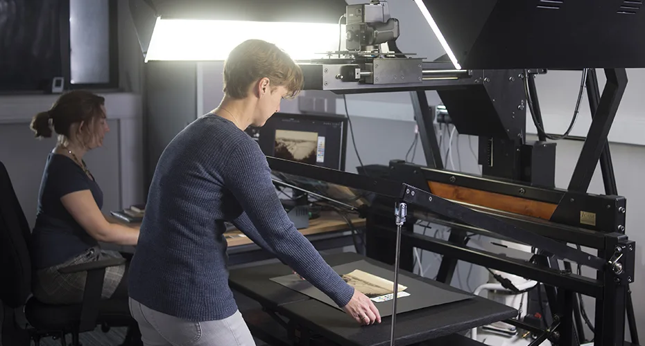 An image of two people using the copystand in Lab 1 to digitise flat material - one adjusts the material to make sure it is in the right place, and one uses the computer to check quality of the image once it is taken.