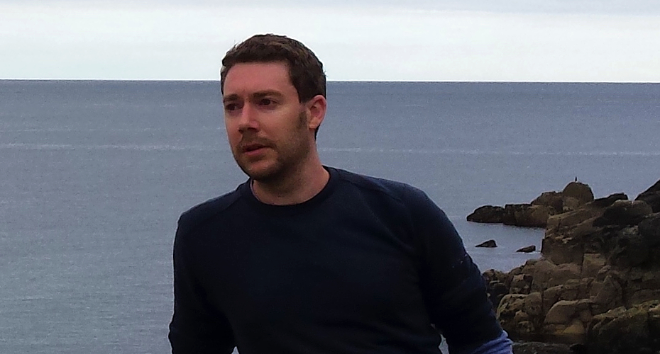 Image of Daniel Partridge sat on a sea wall with the sea behind him, wearing a jumper and looking away from the camera