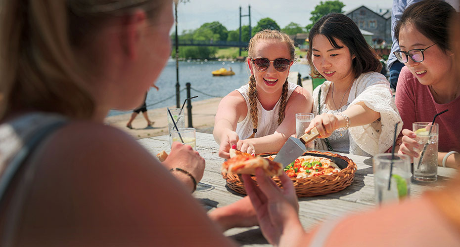 A group of three women savoring pizza together at a table near the water, creating a joyful dining experience.