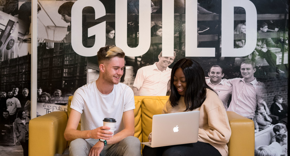 Two individuals seated on a couch, each focused on their laptops, creating a collaborative and relaxed workspace.