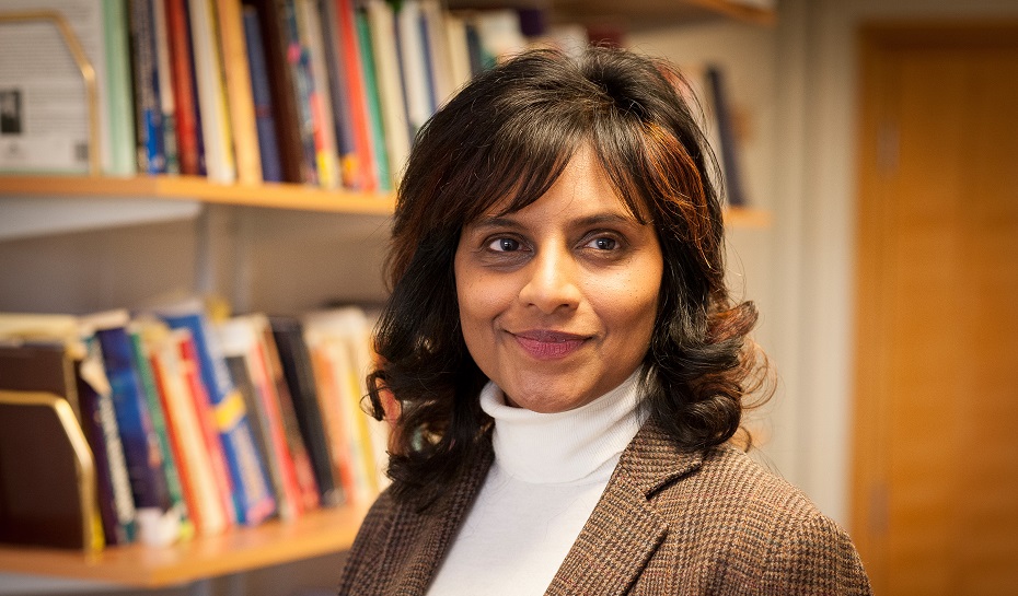 A photograph of Professor Rajani Naidoo. She has dark, shoulder-length hair and is wearing a white polo neck and brown jacket. Behind her is a series of bookshelves lined with various books.