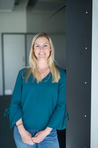 Jen mcwhorter, dressed in a blue shirt and jeans, stands confidently in a contemporary office space.