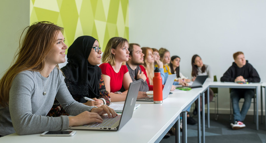 A diverse group of students collaborates at tables, each using laptops in a vibrant lecture space.